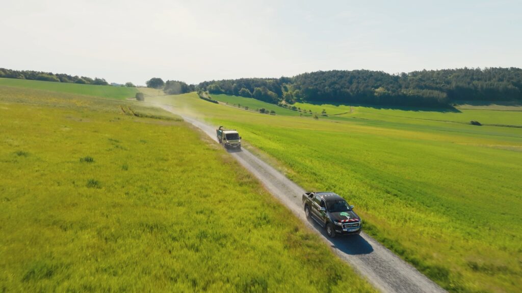 Drohnenaufnahme-feldweg-nutzfahrzeuge-pickup-lkw-grüne-felder-staubspur-outdoor-imagefilm-dreh-logistik-transport-b2b