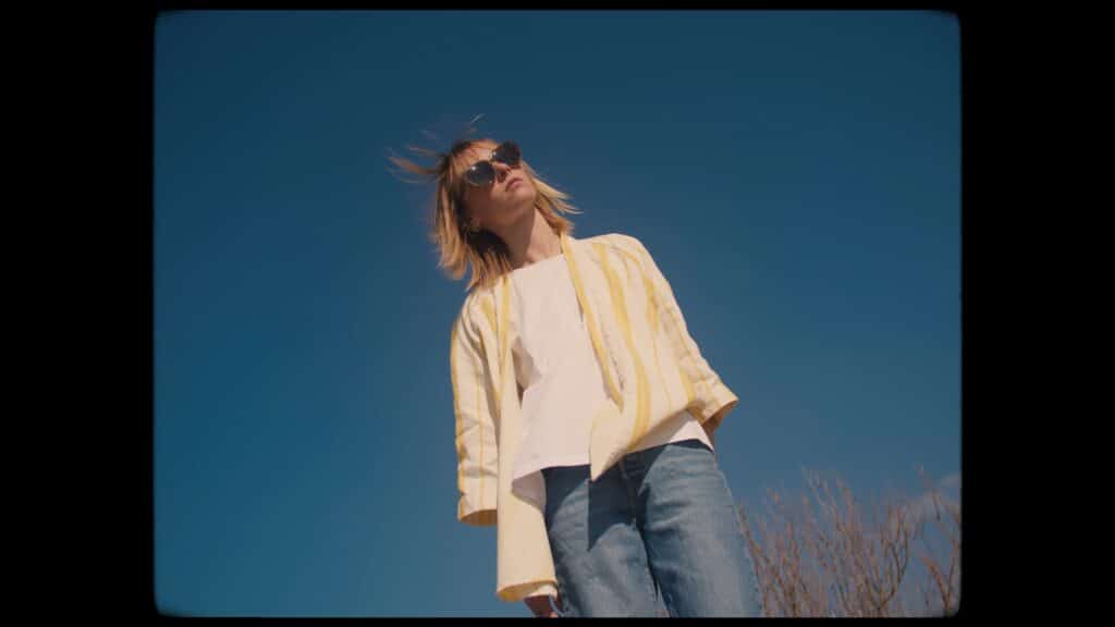 Imagefilm-Dreh, Low-Angle-Aufnahme, Outdoor-Szene, Person-mit-Sonnenbrille, Blauer-Himmel, Natürliches-Licht, Cinematic-Look, Wind-im-Haar, Weitwinkel-Perspektive, Ästhetische-Farbbearbeitung