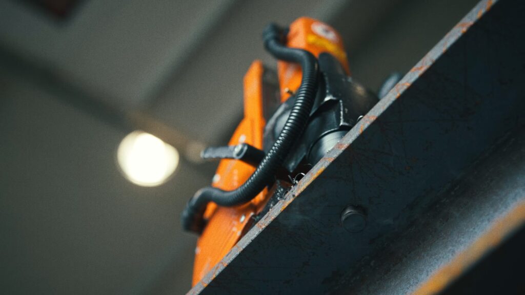 industrieumgebung-nahaufnahme maschinen-detail produktvideo-fokus stahlträger-struktur orange-arbeitsmaschine hydraulik-schläuche cinematic-makroaufnahme fabrikhalle-bokeh
