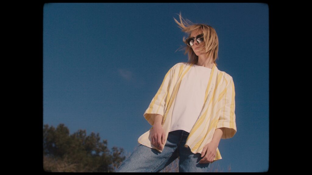 Imagefilm-Dreh, Lifestyle-Outdoor, Low-Angle-Perspektive, Blauer-Himmel, Sonnenbrillen-Detail, Wind-im-Haar, Cinematic-Look, Natürliche-Lichtstimmung, Marken-Inszenierung, Farbbearbeitung-Optimierung