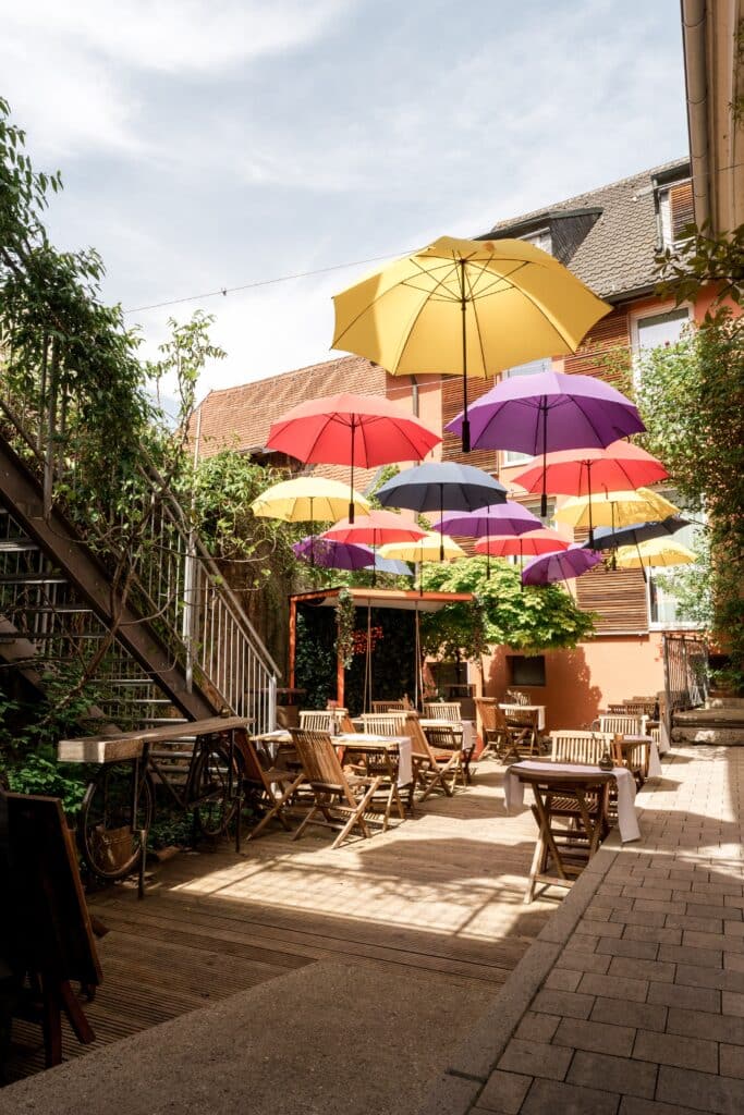 innenhof-cafe-terrasse, hängende-regenschirme, bunte-deko, sommerliches-available-light, location-scouting, imagefilm-dreh, b-roll-gastronomie, architektur-details, outdoor-interview-setup-potenzial