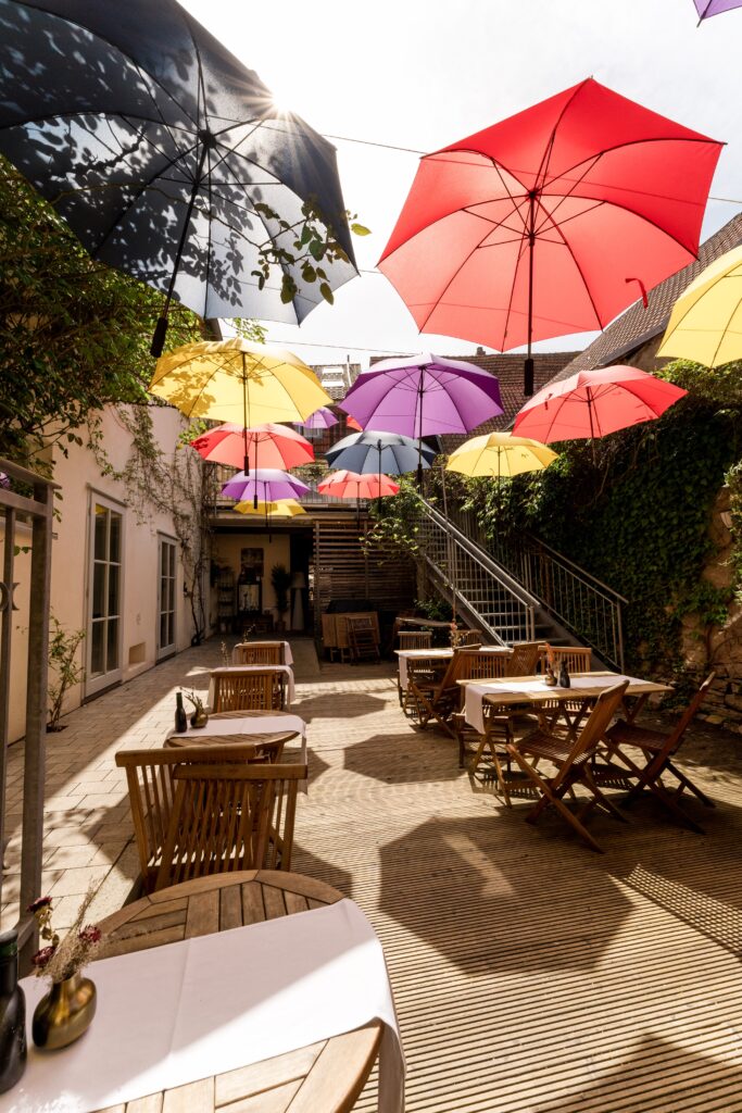 Innenhof-Terrasse Gastronomie-Location Bunte-Schirme Sommerliches-Tageslicht Imagefilm-Dreh B-Roll-Aufnahmen Outdoor-Set Holz-Mobiliar Schatten-Spiel Urbaner-Charme