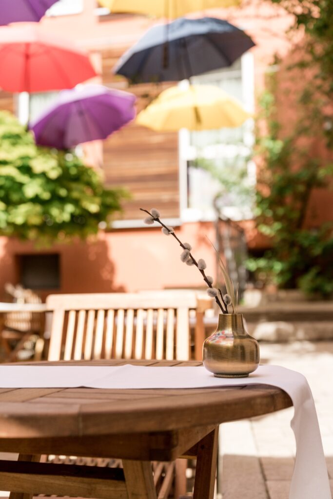 imagefilm-dreh, outdoor-location, gastro-terrasse, holz-tisch-dekor, messing-vase, kätzchen-zweig, bunte-schirme, natürliches-licht, detail-aufnahme, b-roll, lifestyle-setting