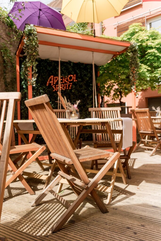 Außenbereich-Gastronomie, Gastro-Terrasse, Holzmöbel-Setup, Sonnenschirm-Atmosphäre, Branding-Neonschild, Tischdeko-Detail, Sommer-Location, Imagefilm-Dreh, Produktvideo-Getränke, Lifestyle-Szene, Available-Light-Aufnahme