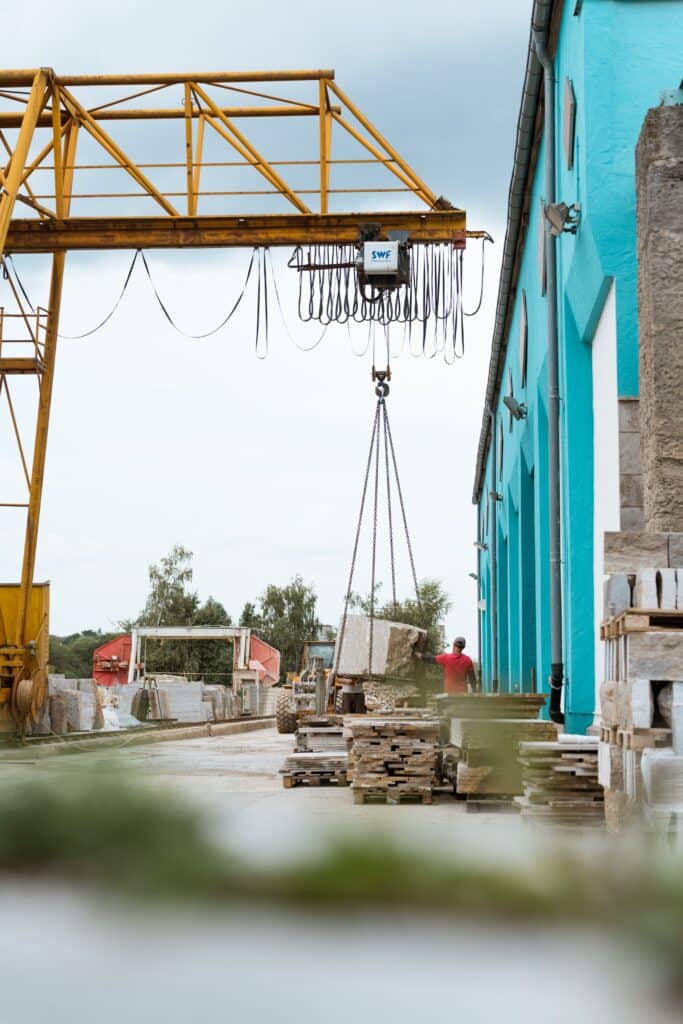 industrieumgebung-steinverarbeitung-hallenkran-hubvorgang-natursteinblock-lagerplatz-paletten-werkshof-mitarbeiter-sicherer-umschlag-prozessaufnahme-imagefilm-dreh-b2b-produktvideo-recruiting-video