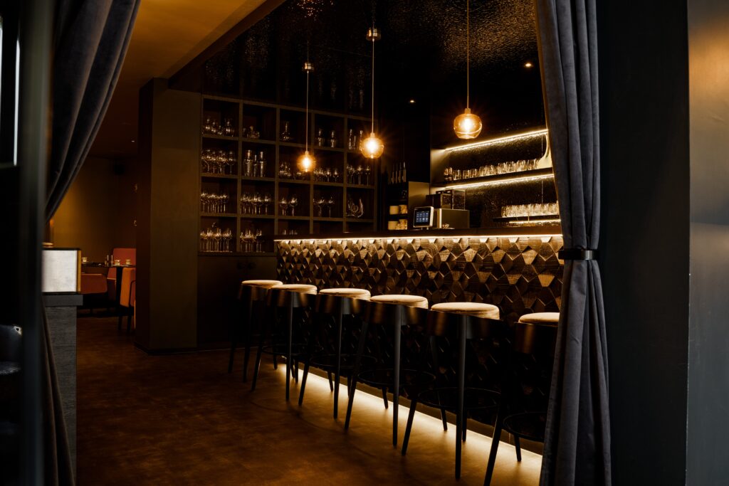 Bar-Interieur, Gastronomie-Location, stimmungsvolle-Beleuchtung, Thekenbereich-Design, Pendelleuchten-Setup, Glasregal-Details, Hochhocker-Reihe, Warmton-Licht, Lowlight-Dreh, Cinematic-Look, Imagefilm-Dreh, B-Roll-Aufnahmen, Produktvideo-Gastronomie, Location-Scouting, Innenaufnahme-Architektur