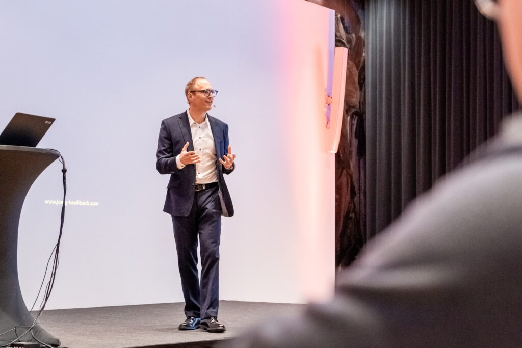 Eventfilm-Keynote, Konferenzfilm-Bühnenvortrag, Corporate-Event, Keynote-Aufzeichnung, Headset-Mikrofon, Präsentations-Setup-Stehpult, Bühnenlicht-LED, Kameraperspektive-Publikum, Business-Storytelling, Livestream-Produktion