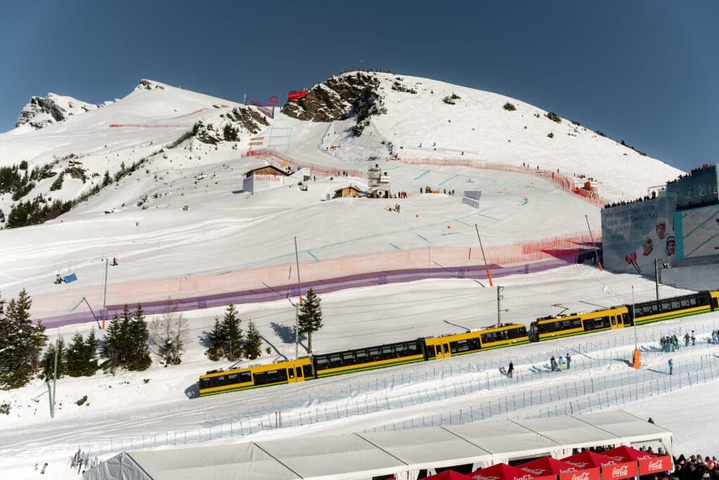 drohnenaufnahme-skirennen alpen-panorama bergbahn-vordergrund pistenabsperrung-netze werbebanden-branding eventproduktion-winter outdoor-sportevent livestream-setup zuschauer-bereich schneesport-location