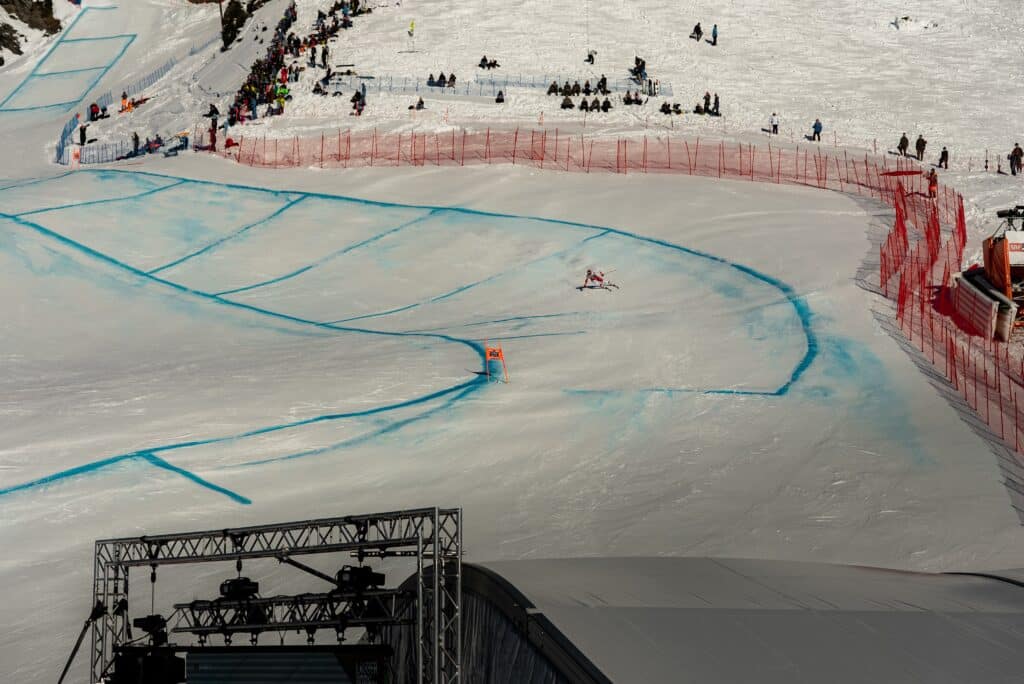 Drohnenaufnahme-Skirennen, Sportevent-Dreh, Outdoor-Winterlandschaft, Pistenkurs-Markierungen, Sicherheitszäune-Zuschauerbereich, Kamera-Rig-Bühnenaufbau, Liveproduktion-Setup, Action-Sequenz
