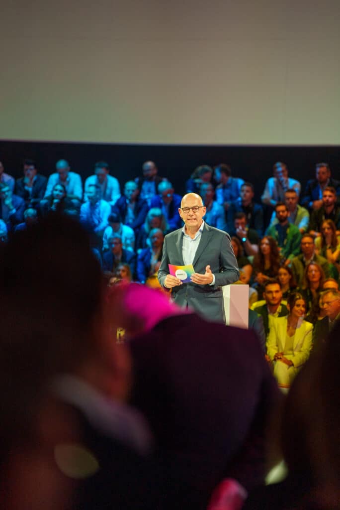 Eventfilm-Dreh, Keynote-Bühne, Business-Konferenz, Live-Moderation, Publikum-im-Hintergrund, Bühnenlicht-Inszenierung, Corporate-Event, Messefilm-Content